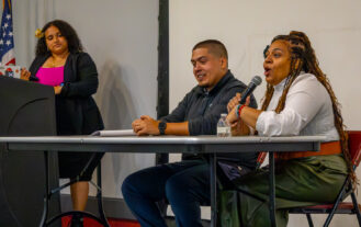 A man and a woman sit at a table on a stage. The woman is speaking into a microphone. Another woman stands at a podium to the left of the table.