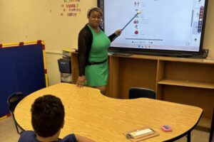 A woman with a teal dress and black cardigan stands at a white board holding a pointer. A boy sits at a table facing the white board.