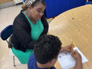 A woman wearing a teal dress and black cardigan sits at a table with a boy while he works on a worksheet.