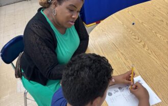A woman wearing a teal dress and black cardigan sits at a table with a boy while he works on a worksheet.