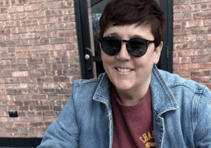 A woman with short dark hair and sunglasses smiles. There is a brick background. 