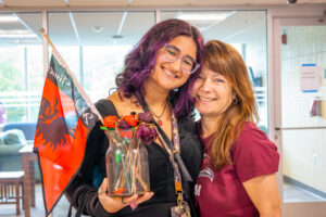 A female college student with purple hair, holding a vase of dried flowers, stands close to a woman with a maroon shirt who is her mother.