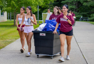 A group of four female college students walk down a path with a large bin on wheels filled with items for a dorm room.