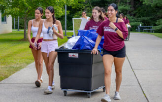A group of four female college students walk down a path with a large bin on wheels filled with items for a dorm room.