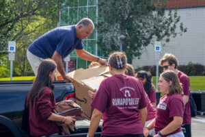 A group of students wearing maroon shirts surround a man standing in a pickup truck as he hands them a large cardboard box.