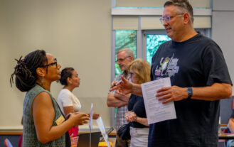 A woman wearing a green vest looks up at and speaks to a tall man wearing a blue T-shirt, holding a piece of paper.