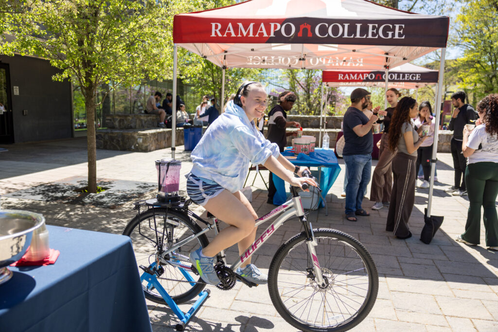 A student pedals a stationary bike with a blender attached to the back. Pedaling powers the blender to make a smoothie at the RCNJ Wellness Fair outside in The Grove on campus.