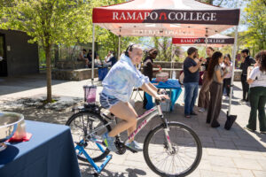 A student pedals a stationary bike with a blender attached to the back. Pedaling powers the blender to make a smoothie at the RCNJ Wellness Fair outside in The Grove on campus.