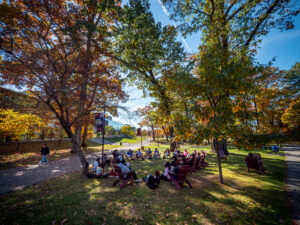 A class sits in a circle outside in the Grove, taking advantage of the fall weather under the canopy of trees with colorful foliage on a clear blue sky day.