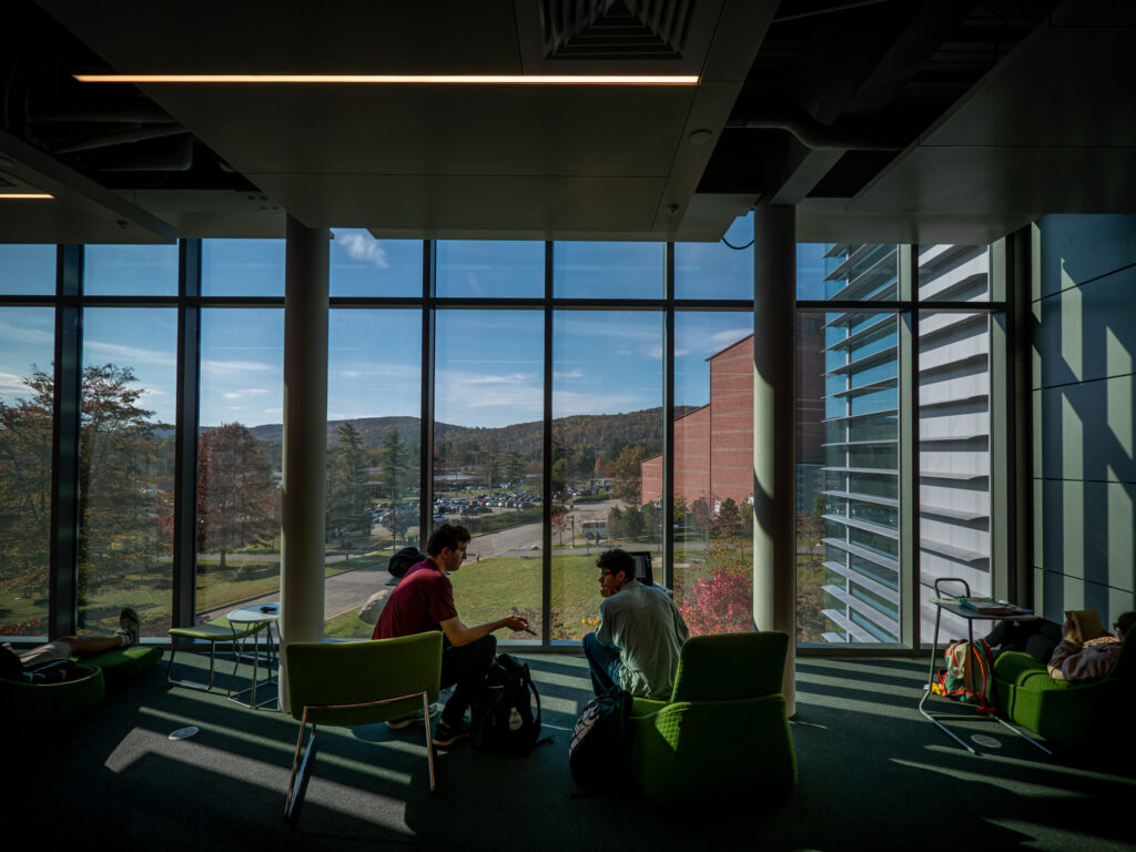Two students sit on chairs in front of floor-to-ceiling windows inside the Peter P. Mercer Learning Commons. The view is of the Berrie Center and the Ramapo Mountains in the far background.
