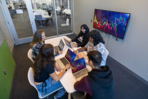A group of five college students sit around a table, each with a laptop open in front of them. They are in a small meeting room with glass doors, and a monitor hanging on the wall.