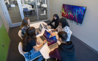 A group of five college students sit around a table, each with a laptop open in front of them. They are in a small meeting room with glass doors, and a monitor hanging on the wall.