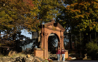 Two students in Ramapo College shirts walk toward the camera from the Havemeyer Arch on a fall day. The trees are covered in green, orange, and red leaves.