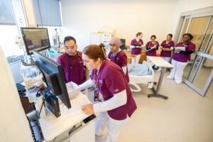 Two nursing students in maroon and white scrubs stand around a monitor, while more students stand behind them in a simulation lab that looks like a hospital room.