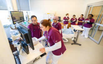 Two nursing students in maroon and white scrubs stand around a monitor, while more students stand behind them in a simulation lab that looks like a hospital room.