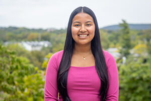 A woman with long dark hair and a pink shirt stands and smiles with the mountains behind her.
