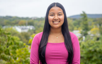 A woman with long dark hair and a pink shirt stands and smiles with the mountains behind her.