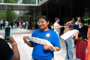 A female college student in a blue T-shirt stands outside and holds a roll of voting stickers in front of her, as someone takes her picture with a cell phone.