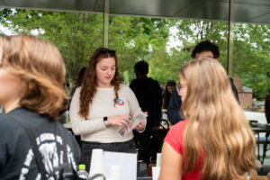 A female college student stands behind a table while two students stand in front of it with their backs to the camera.