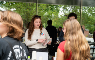 A female college student stands behind a table while two students stand in front of it with their backs to the camera.
