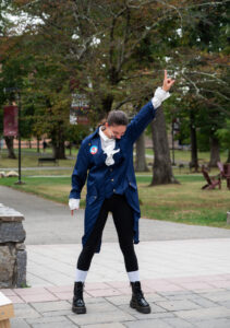 A college student standing up outside is dressed as Alexander Hamilton. She is standing up, with her head down, and one hand pointed in the air.