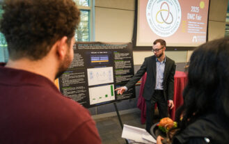 A student wearing a suit points to a research poster and speaks to guests about his research. Behind him is a projector screen displaying the DMC Center logo with the words 2025 Fair.