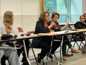Four people sit behind a table. One has a microphone and speaks as the others listen.