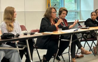 Four people sit behind a table. One has a microphone and speaks as the others listen.
