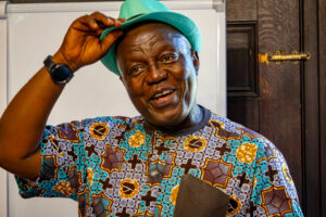 A man smiles while he holds the edge of his teal fedora on top of his head.