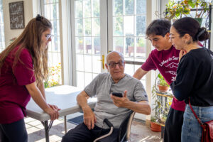 Three college students stand around an older gentleman sitting down and holding a phone.