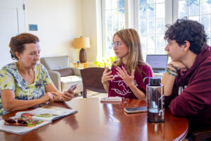 An older woman sits at a table with two college students wearing maroon shirts.