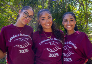Three smiling young women with maroon shirts and stand shoulder to shoulder outside.