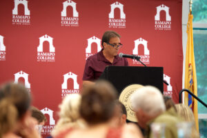 A man with a maroon shirt stands behind a podium and speaks to a crowd. The backs of a few heads can be seen in front of him.