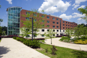 An exterior photo of Mackin Hall, a first-year residence hall, shows the side facade of the building. It has four stories and a fully-windowed stairwell on the left end of the building. There is a courtyard in the foreground featuring trees, grass, and a circular paved path.