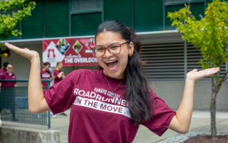 A student with long hair and glasses wears a maroon RCNJ t-shirt that says "Roadrunners on the move" and stands in front of The Overlook, a res hall on campus. She smiles and holds her arms out on each side of her in the air with enthusiasm.