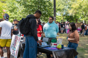 A student stands in front of a table outside, while two more students stand behind the table smiling at him.