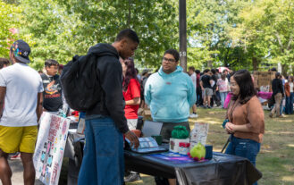 A student stands in front of a table outside, while two more students stand behind the table smiling at him.