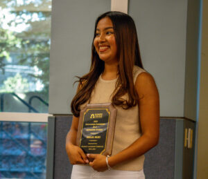 A female college student stands and smiles while holding a plaque.