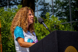 A woman stands at a podium and speaks, with trees behind her.