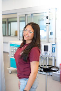 A smiling college student with a maroon shirt stands in front of nursing equipment.