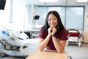 A female college student rests her elbows on a table and smiles in a nursing lab.