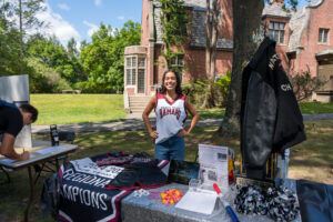 A college student stands under a tree and behind a table, smiling.