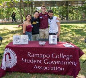 A group of four students stand behind a table with a maroon table clothes outside.