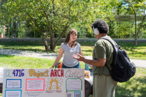 A college student with a white shirt stands behind a poster board and speaks to another student in green.