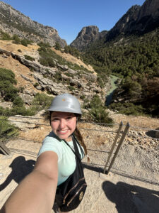 A young woman wearing a helmet takes a selfie by a river with a mountain in the background.