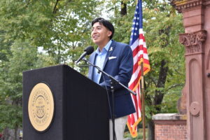 A man in a blazer speaks at a podium outside. The American flag is behind him.
