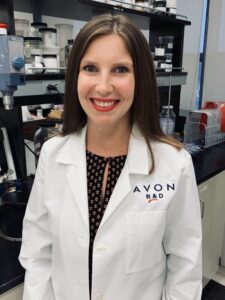 A woman wearing a white lab coat stands in a lab and smiles.