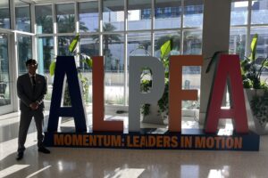A male college student in a gray suit stands in a building lobby next to large letters ALPFA.