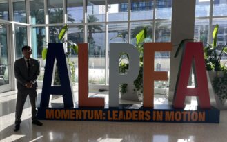 A male college student in a gray suit stands in a building lobby next to large letters ALPFA.