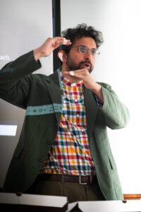 A man with a green blazer stands and talks in front of a whiteboard, holding his hands up like he is measuring something.